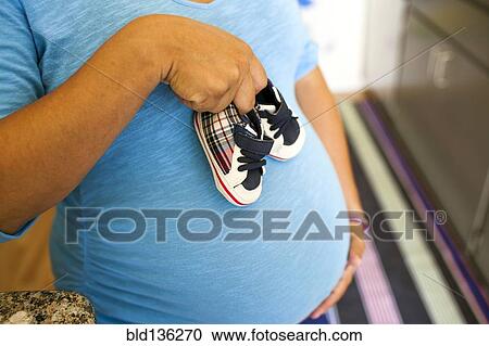 Stock Image - Pregnant Hispanic woman holding baby shoes. Fotosearch