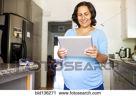 Stock Image - Pregnant Hispanic woman using tablet computer in kitchen. Fotosearch