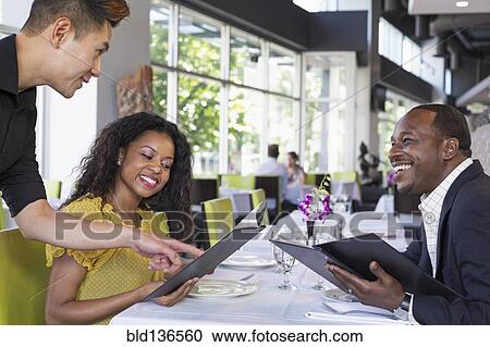 Stock Image - Waiter explaining menu to couple in restaurant. Fotosearch