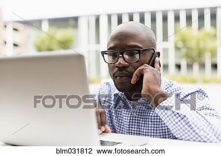 Stock Image - Black man using cell phone and laptop outdoors. Fotosearch