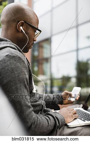 Stock Photography - Black man using laptop, earphones and cell phone outdoors. Fotosearch