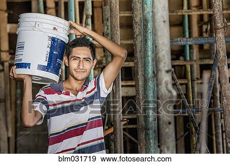 Stock Photo - Hispanic construction worker carrying bucket at construction site. Fotosearch