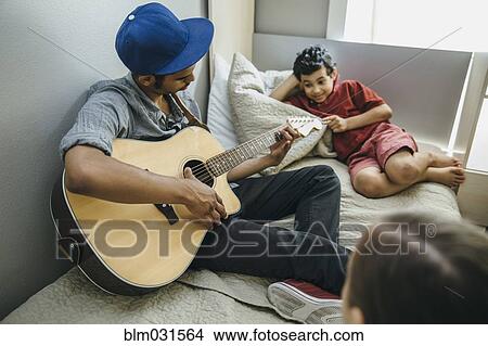 Picture - Mixed race boy watching brother playing guitar in bedroom. Fotosearch