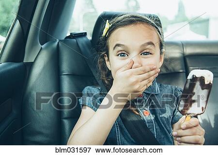 Stock Photo - Mixed race girl eating ice cream bar in car back seat. Fotosearch