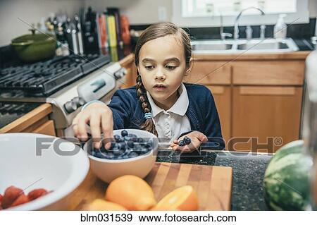 Stock Photo - Mixed race girl picking blueberries from bowl in kitchen. Fotosearch