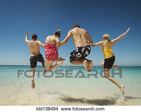 Picture - Caucasian couples jumping for joy in water on tropical beach. Fotosearch