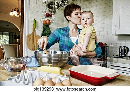 Stock Image - Caucasian mother kissing baby and cooking in kitchen. Fotosearch