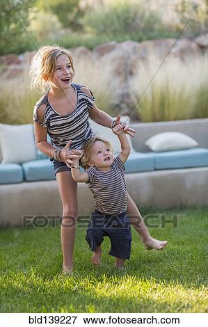 Stock Photo - Caucasian sister helping toddler brother walk in backyard. Fotosearch