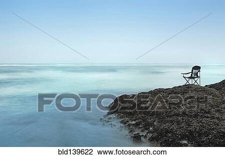 Stock Image - Chair overlooking waves on rocky beach. Fotosearch