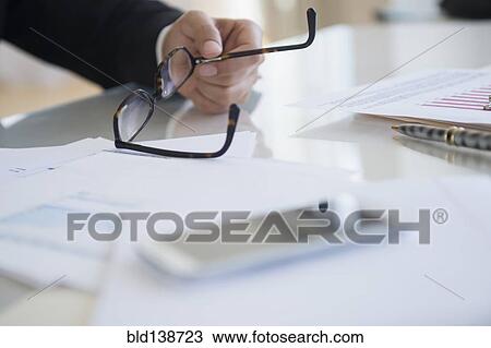 Stock Image - Close up of mixed race businessman holding eyeglasses at desk. Fotosearch