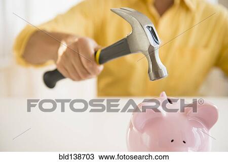 Stock Image - Close up of mixed race man holding hammer over piggy bank. Fotosearch