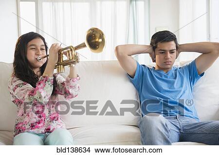 Hispanic brother covering his ears as sister practices trumpet in living room View Large Photo Image Picture - Hispanic brother covering his ears as sister practices trumpet in living room. Fotosearch