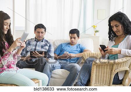 Stock Image - Hispanic family using cell phones and digital tablets in living room. Fotosearch