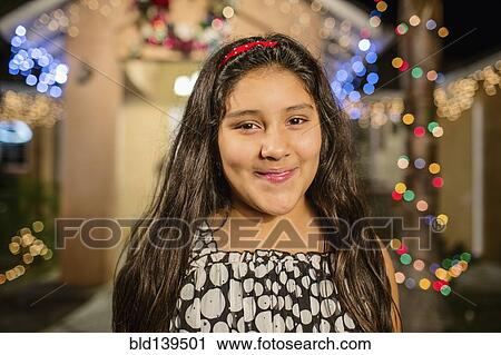 Stock Image - Hispanic girl smiling outside house decorated with string lights. Fotosearch