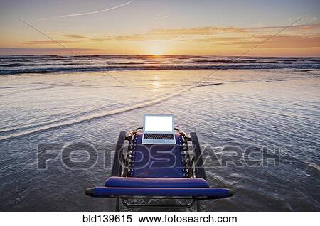 Stock Photography - Laptop on deck chair overlooking sunset on beach. Fotosearch
