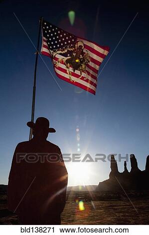 Stock Image - Man holding United States flag with Native American depiction near rock formations, Monument Valley, Utah, United States. Fotosearch