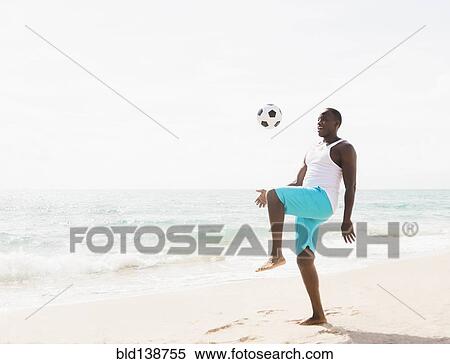 Stock Photography - Mixed race man playing with soccer ball on beach. Fotosearch
