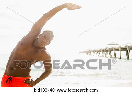 Stock Image - Mixed race man stretching on beach. Fotosearch