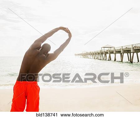 Stock Image - Mixed race man stretching on beach. Fotosearch