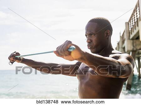 Stock Photo - Mixed race man using resistance band on beach. Fotosearch