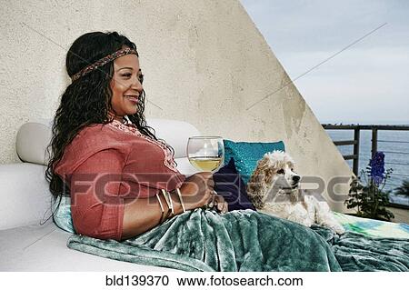 Stock Image - Mixed race woman drinking wine at waterfront with dog. Fotosearch