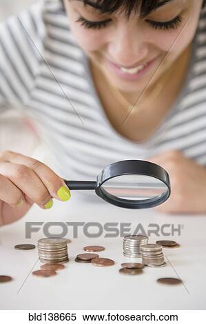 Stock Photography - Mixed race woman examining stacks of coins with magnifying glass. Fotosearch