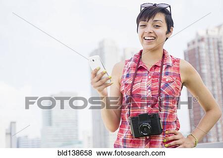Stock Photo - Mixed race woman with camera and cell phone smiling near city skyline. Fotosearch