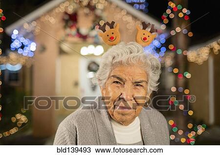 Stock Image - Older Hispanic woman smiling outside house decorated with string lights. Fotosearch