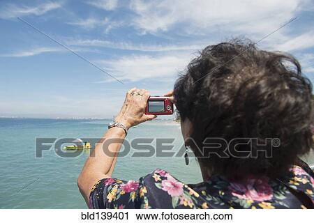 Stock Image - Older woman taking photograph of tropical ocean. Fotosearch
