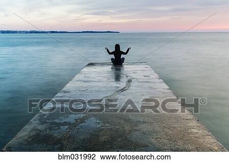 Stock Image - Asian woman meditating on concrete pier. Fotosearch