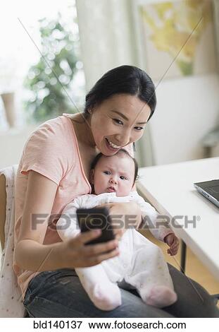 Stock Photo - Asian mother and baby taking selfie in home office. Fotosearch