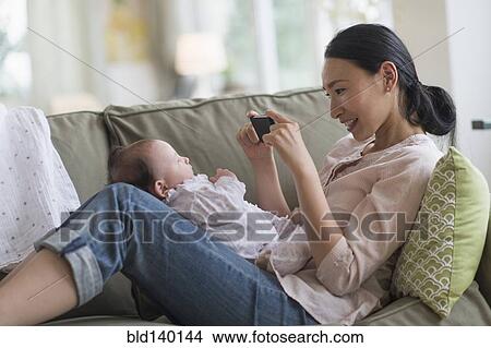 Picture - Asian mother taking cell phone photograph of baby in living room. Fotosearch