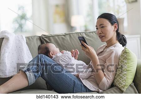 Stock Image - Asian mother using cell phone and holding baby in living room. Fotosearch