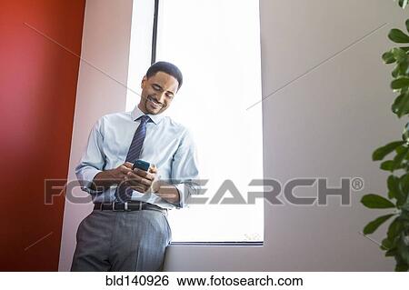 Stock Photograph - Black businessman using cell phone at office window. Fotosearch