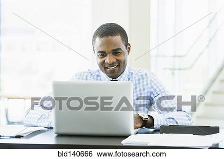 Stock Photograph - Black businessman using laptop computer at desk in office. Fotosearch