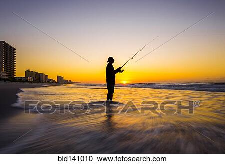 Stock Image - Blurred view of silhouette of man fishing in waves on beach at sunset. Fotosearch