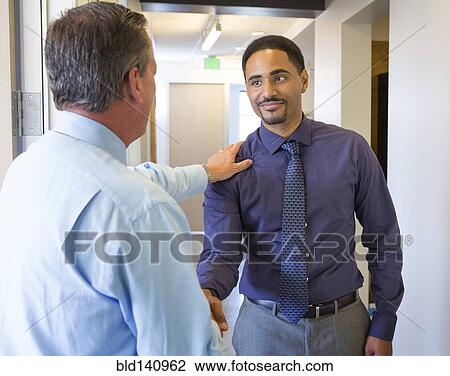 Stock Image - Businessmen shaking hands in office hallway. Fotosearch