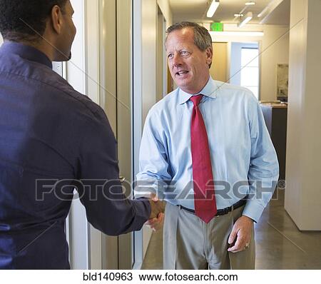 Stock Image - Businessmen shaking hands in office hallway. Fotosearch
