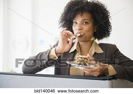 Stock Photograph - Businesswoman eating stack of cookies in cubicle. Fotosearch