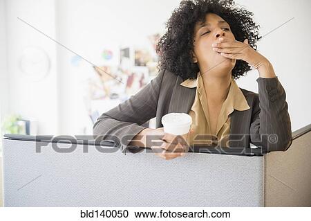 Stock Image - Businesswoman with cup of coffee yawning in cubicle. Fotosearch