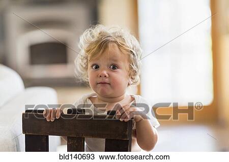 Stock Image - Caucasian baby boy standing behind chair in living room. Fotosearch
