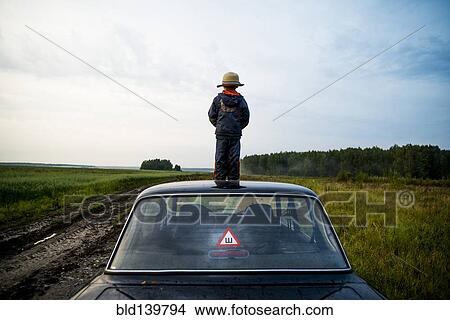 Picture - Caucasian boy standing on car roof in rural field. Fotosearch