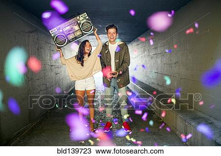 Stock Image - Caucasian couple carrying boom box and throwing confetti in tunnel. Fotosearch