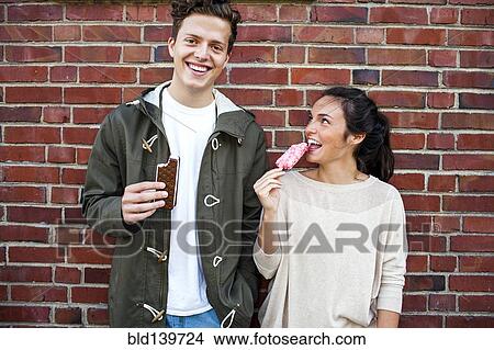 Picture - Caucasian couple eating ice cream near red brick wall. Fotosearch