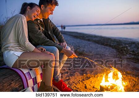 Stock Image - Caucasian couple roasting marshmallows on fire at beach. Fotosearch