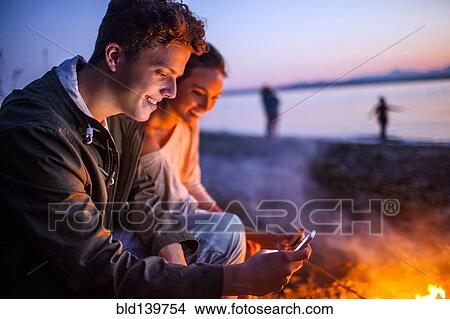 Picture - Caucasian couple using cell phone together near fire on beach. Fotosearch