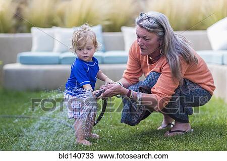 Stock Image - Caucasian grandmother and baby grandson watering lawn with hose. Fotosearch
