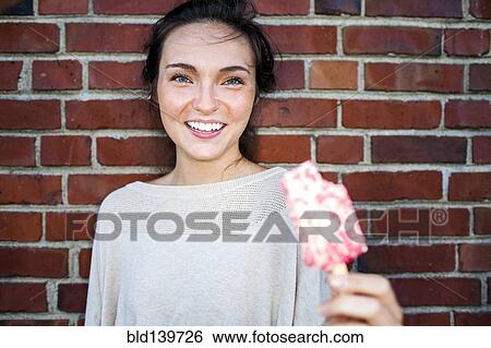Stock Photograph - Caucasian woman eating ice cream near red brick wall. Fotosearch