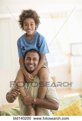 Stock Image - Father carrying son on shoulders in living room. Fotosearch