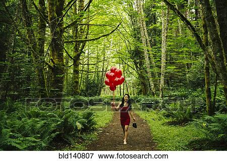 Stock Photo - Korean woman holding red balloons in lush forest. Fotosearch